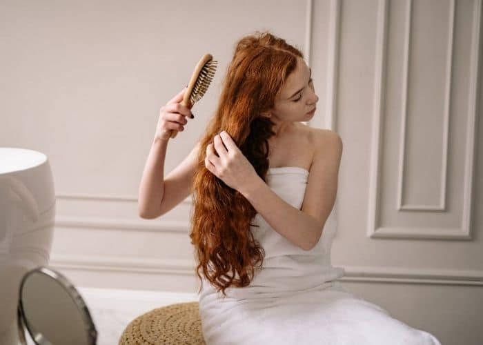 A woman wrapped with a towel brushes her long auburn hair extensions with a wide-tooth hair brush in a living room as a good hair extension care practice
