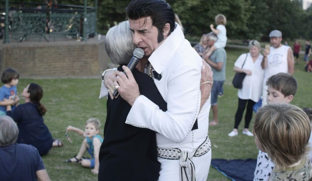 Elvis Presley dressed up in his Elvis hair hugging an older lady in a sports field