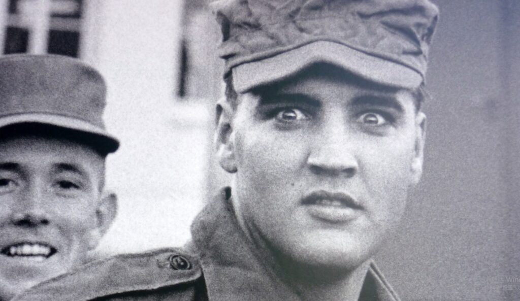 Elvis Presley wearing an army hat, covering his Elvis hair, with his mate in the background